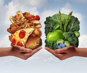 Nutrition choice and diet decision concept and eating choices dilemma between healthy good fresh fruit and vegetables or greasy cholesterol rich fast food with two hands holding food trying to decide what to eat.